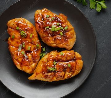 Three grilled chicken breasts topped with teriyaki sauce and a hint of sweet local honey, garnished with chopped green onions and parsley on a black plate, set against a dark background.