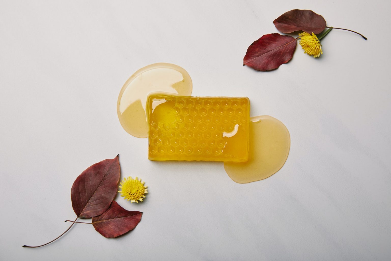 A bar of honey soap on a light surface with sweet local honey drips, red leaves, and yellow flowers arranged around it.