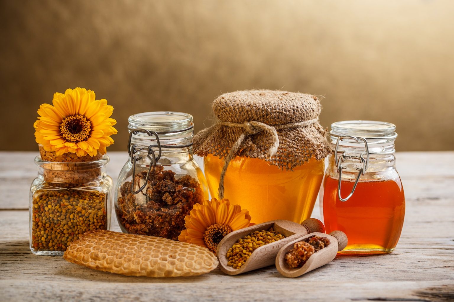 Jars of sweet local honey and honeycombs with yellow flowers, bee pollen, and propolis are arranged neatly on a wooden surface.