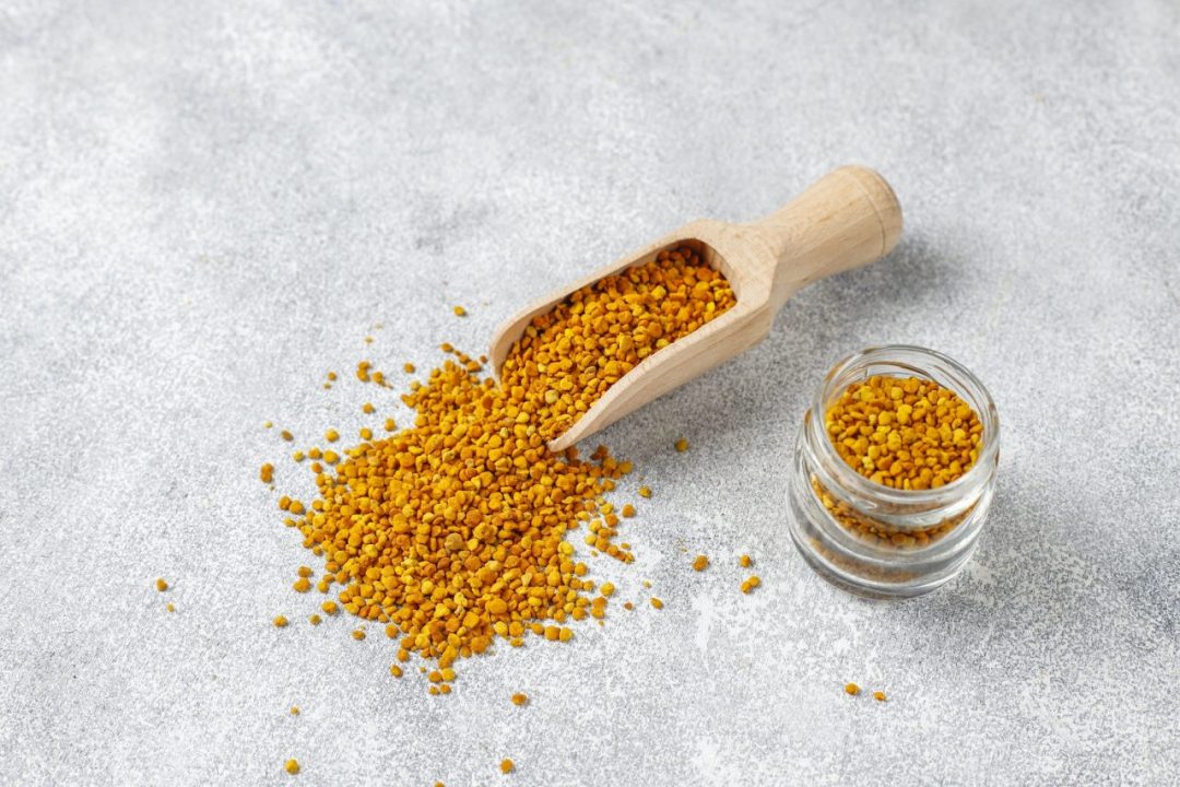 A wooden scoop pours out yellow bee pollen granules next to a small glass jar filled with more of the granules, set on a grey textured surface, accompanied by a drizzle of sweet local honey.