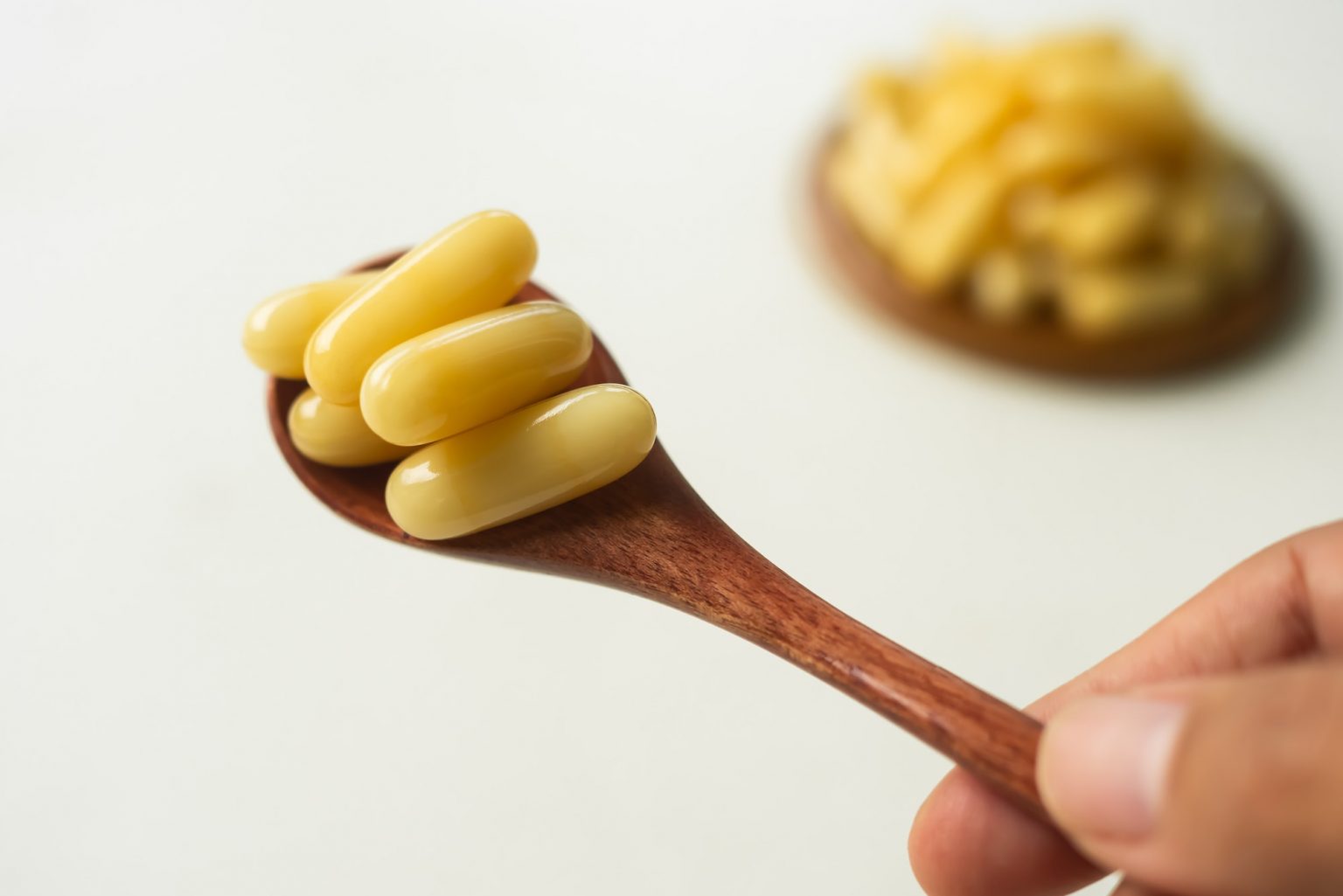 A hand holds a wooden spoon with several yellow capsules, reminiscent of sweet local honey. In the blurred background, there is a wooden plate with more capsules.
