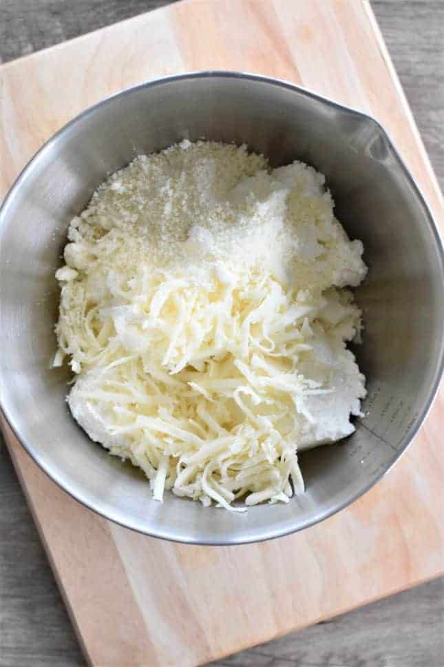 A metal mixing bowl containing shredded cheese, ricotta cheese, and a drizzle of sweet local honey sits on a wooden cutting board. A metal mixing bowl containing shredded cheese, ricotta cheese, and a drizzle of sweet local honey sits on a wooden cutting board.