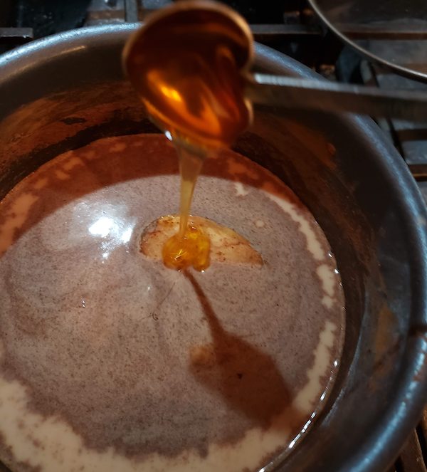 A spoon is pouring sweet local honey into a pot containing a mixture of liquid and a dollop of creamy substance, on a stove. A spoon is pouring sweet local honey into a pot containing a mixture of liquid and a dollop of creamy substance, on a stove.