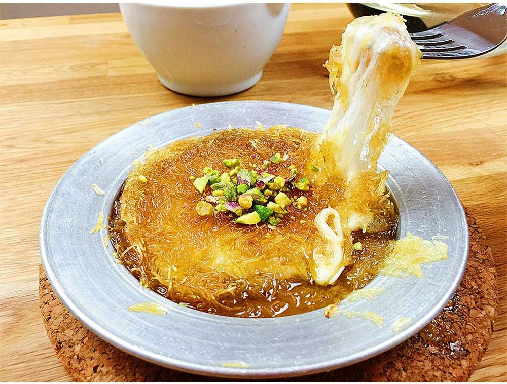 A plate of kunafa, a Middle Eastern dessert with a stretchy cheese filling, topped with chopped pistachios and drizzled with sweet local honey, sits enticingly on a wooden table. A white cup is in the background.