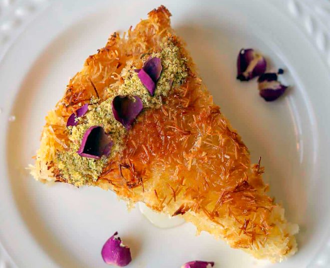 A slice of dessert with a crispy topping, drizzled with sweet local honey, and sprinkled with crushed green pistachios and dried purple flower petals on a white plate. A slice of dessert with a crispy topping, drizzled with sweet local honey, and sprinkled with crushed green pistachios and dried purple flower petals on a white plate.